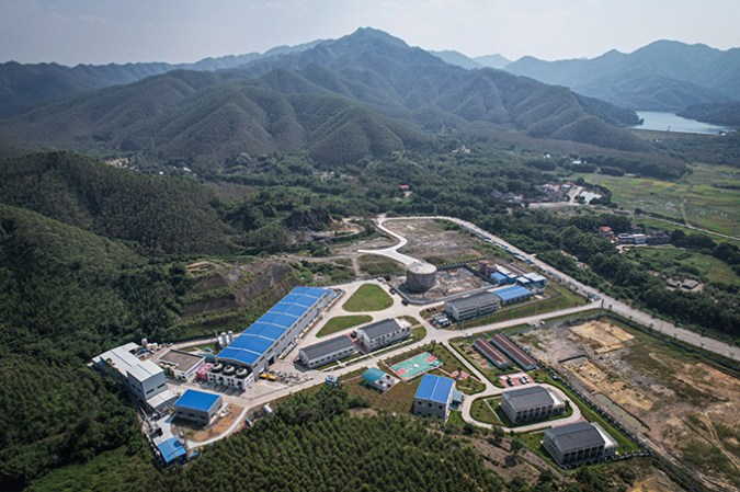 121624_ec_neutrino-detector_slide1-3 A view from above shows laboratory buildings and roadways surrounded by forested mountains.
